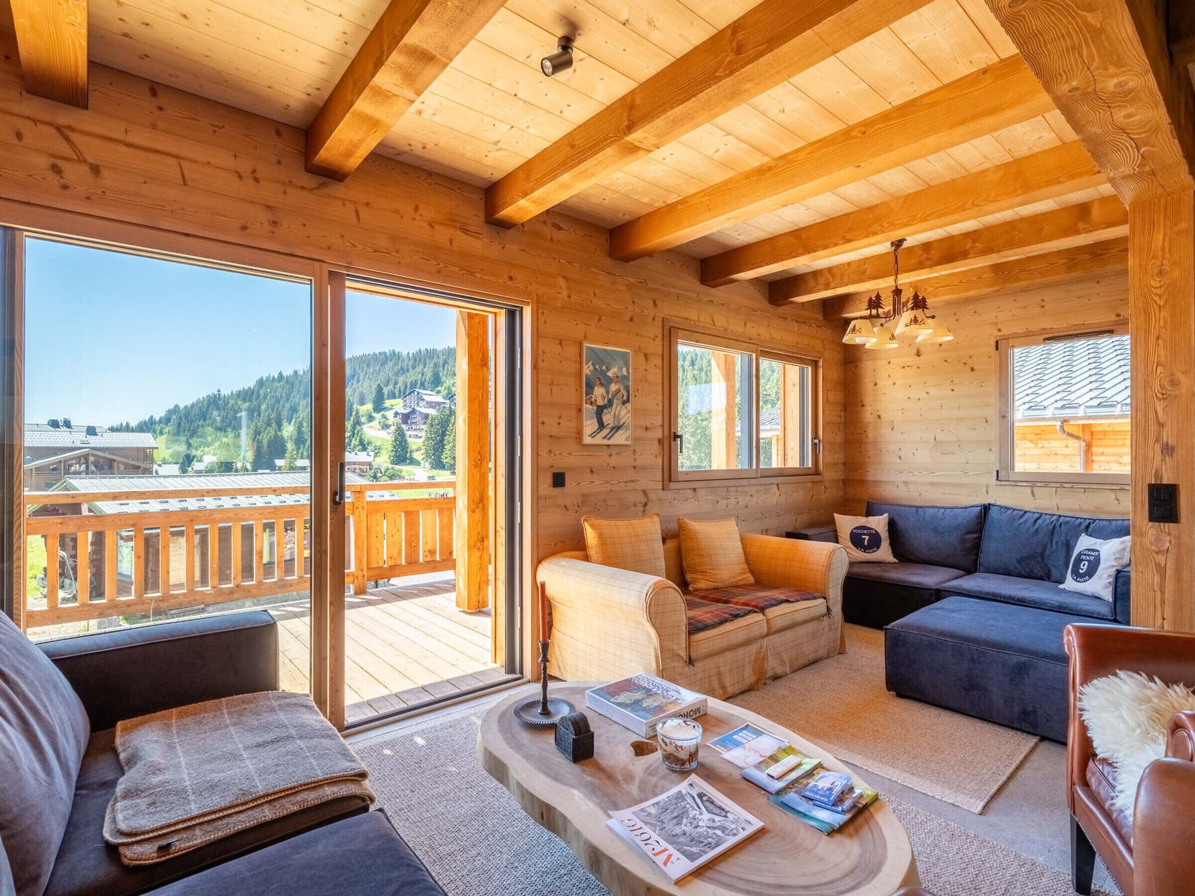 L’intérieur d’un chalet en bois, avec un balcon, un canapé, une table basse avec des livres, et une vue sur un paysage montagneux.
