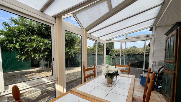 A conservatory with a tiled floor, wooden chairs, a table with a vase of flowers, and a view of a garden.