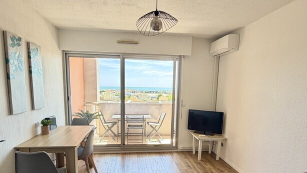 A living room with a sofa, a TV, a dining table with chairs, and a balcony with a view. - 19 STUDIO CLIM VUE MER (Sète)