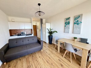 A modern living space with a grey sofa, wooden dining table, and chairs, a kitchen in the background, and a potted plant. - 19 STUDIO CLIM VUE MER (Sète)