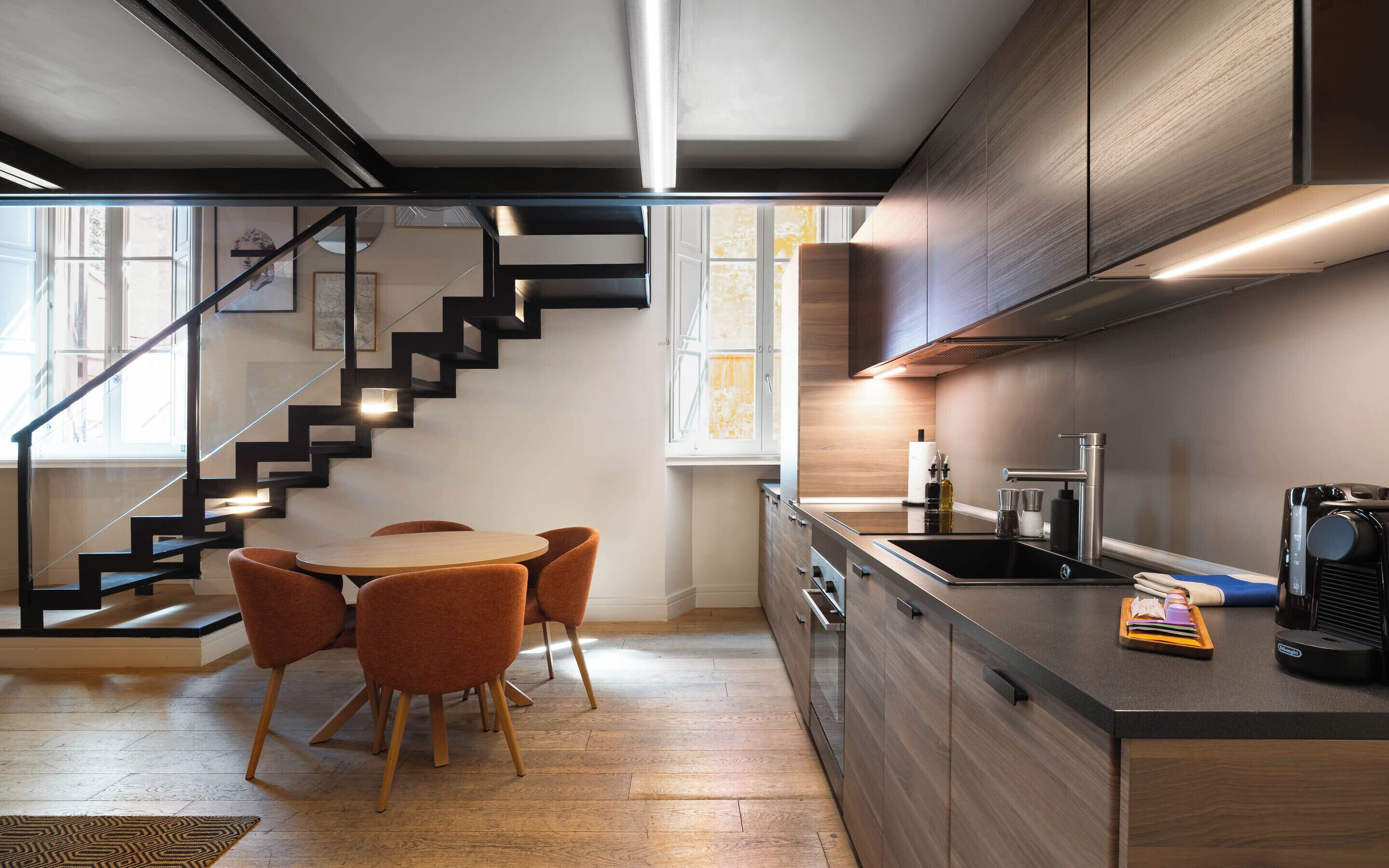 A modern kitchen with a built-in stove, a sink, and a coffee machine. There is a dining area with a table and chairs. A staircase is visible leading to an upper level.
