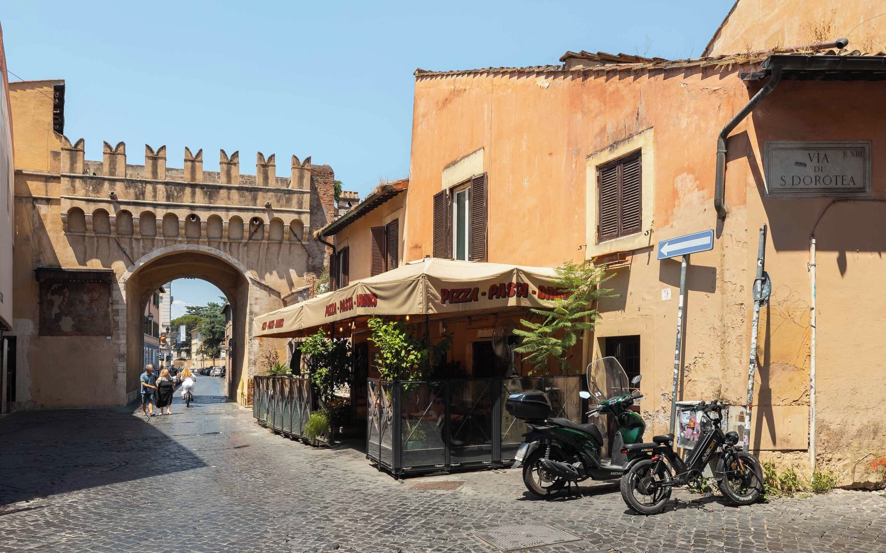 Une rue pavée, ornée d’une arche historique, un restaurant avec une terrasse, et des motos garées.