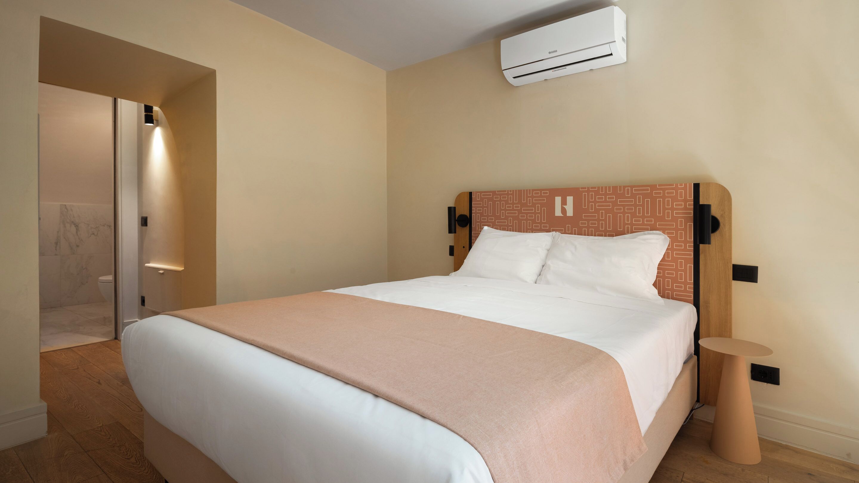 A hotel room with a bed, a bedside table, an air conditioning unit, and a bathroom with a shower.