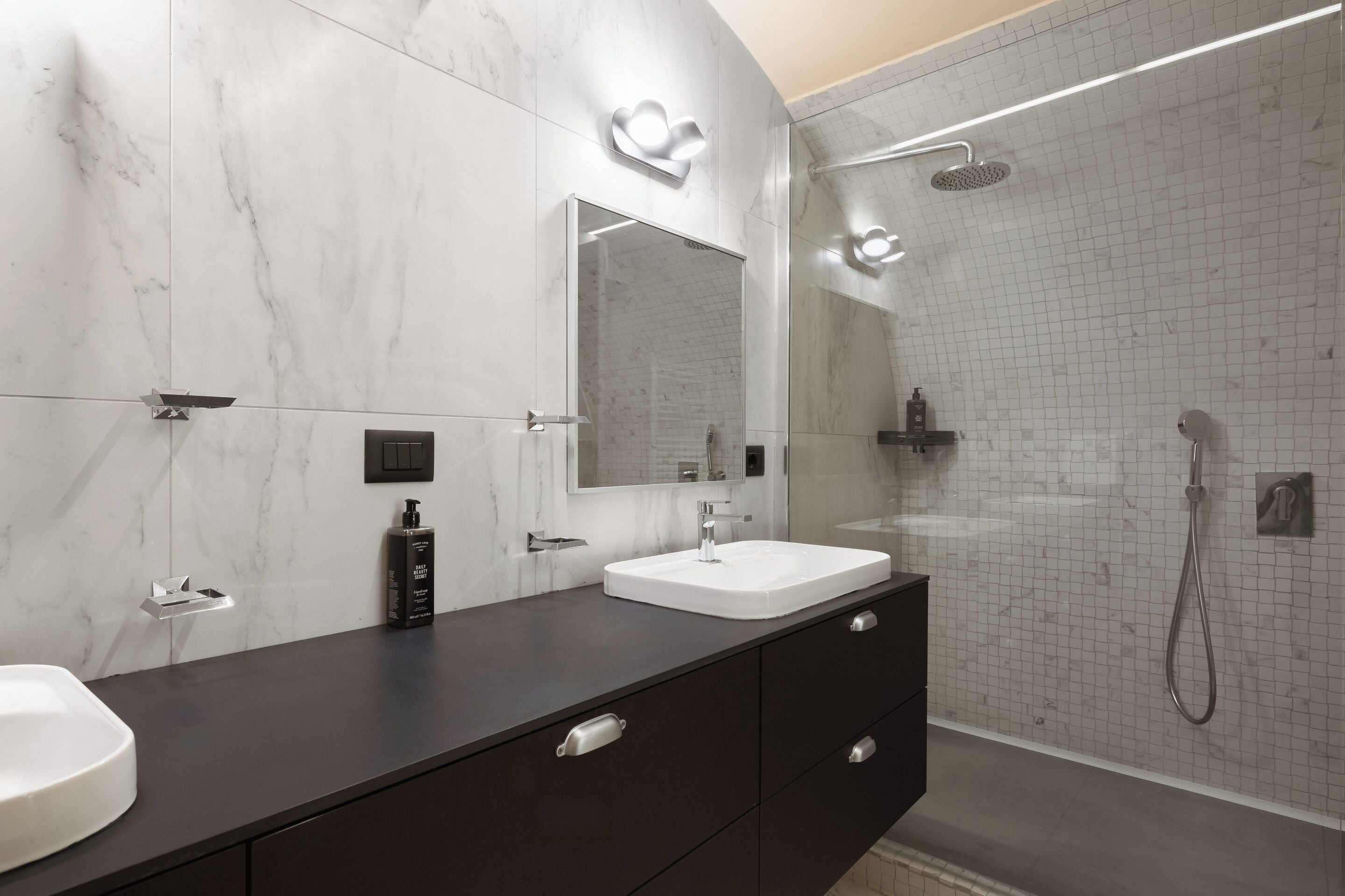 A modern bathroom with a marble wall, a large mirror, a white sink, a dark vanity, and a glass-enclosed shower area.