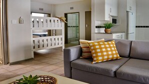 A living room with a grey sofa, a white bunk bed, a kitchen area with white cabinets, and a small plant on the counter. - Kridzil Holiday Flats, Margate, 1-bed Wbt Rentals (Margate)