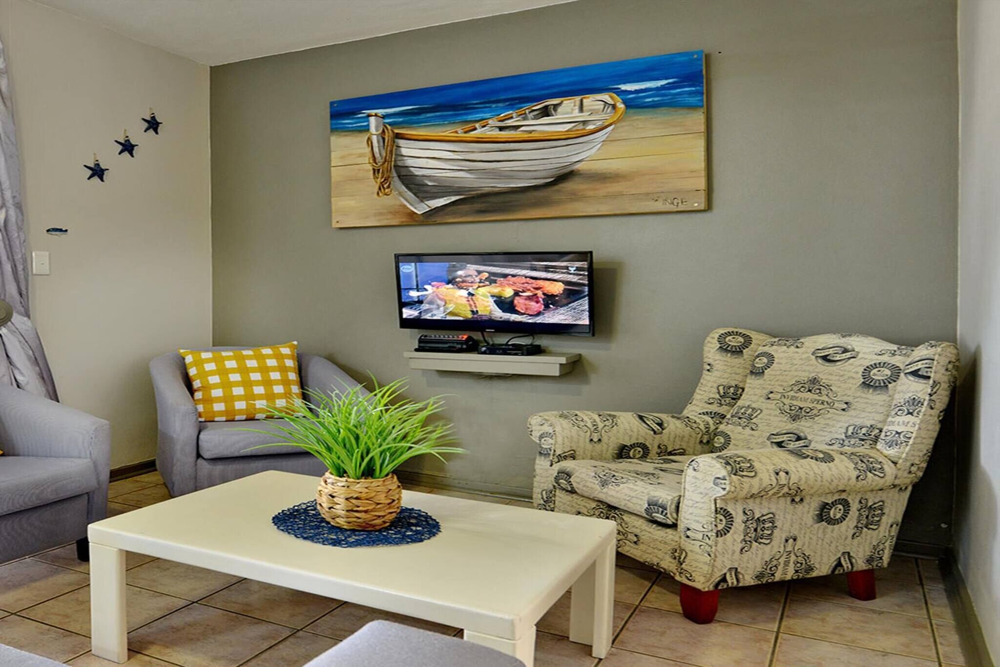 A living room with a television, a patterned armchair, a sofa with a checkered pillow, and a wall-mounted painting of a boat.