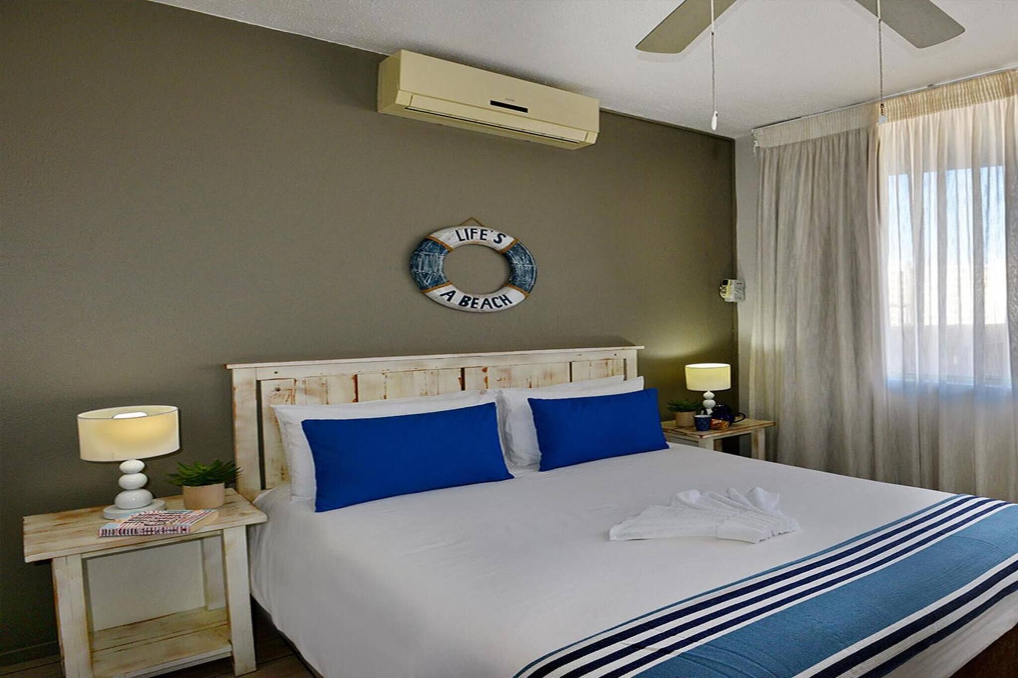 Quarto de hotel com cama, duas mesas de cabeceira, ar-condicionado fixado na parede, um adorno redondo na parede e uma janela com cortinas.