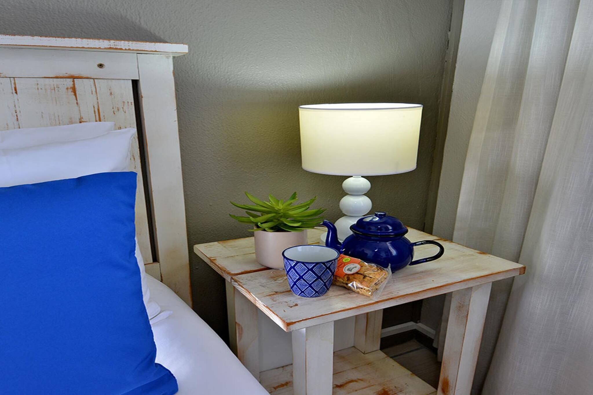 A small bedside table with a lamp, a mug, a teapot, a plant, and a snack pack.