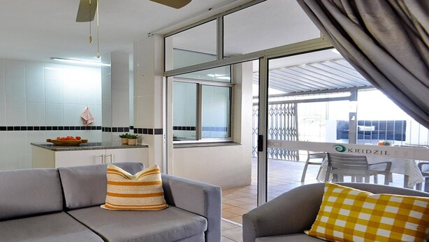 A modern interior with a grey sofa, a checkered pillow, a kitchen area with a tiled backsplash, and a view through a glass door. - Kridzil Holiday Flats, Margate, 1-bed Wbt Rentals (Margate)