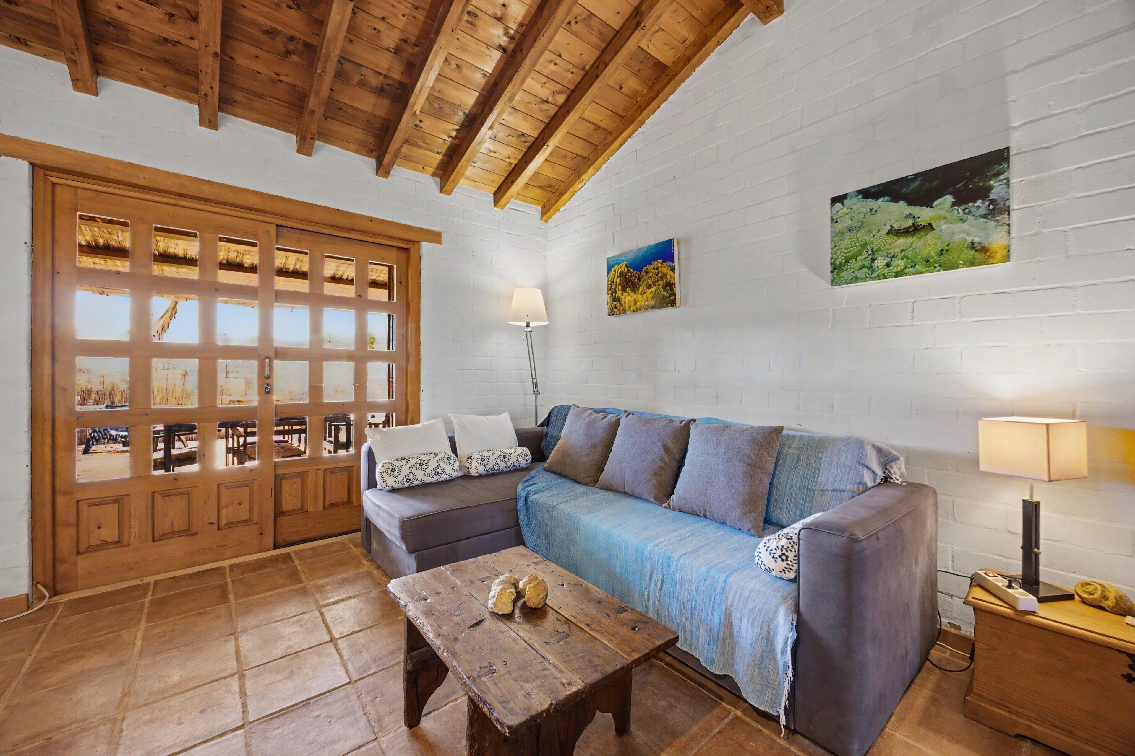 Una habitación con techo de madera, pared de ladrillo blanco, sofá azul, mesa de madera y puerta de madera con paneles de vidrio.