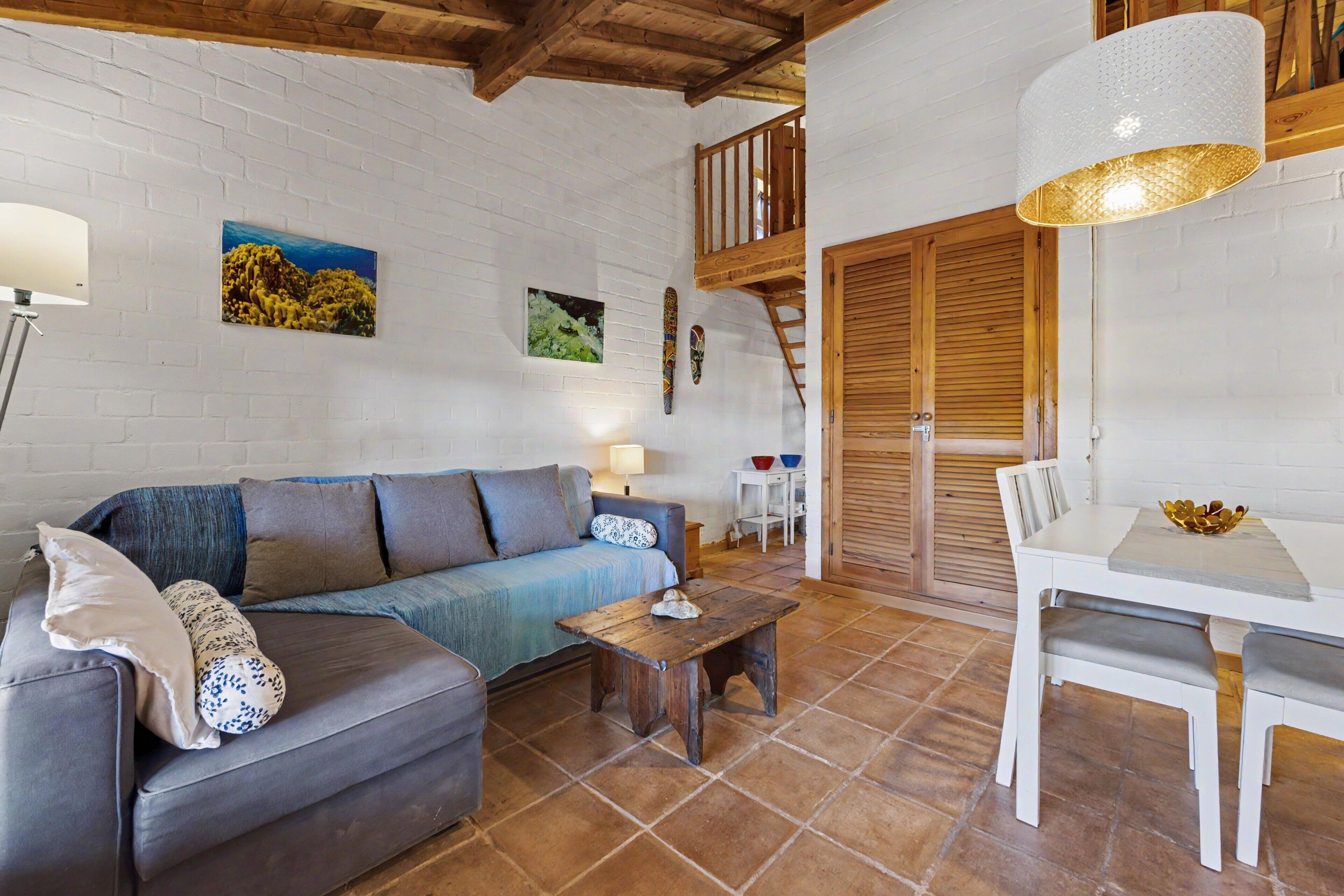 Una sala de estar acogedora con sofá, una mesa de centro y una mesa de comedor con sillas. Se ve una escalera que sube a un nivel superior.