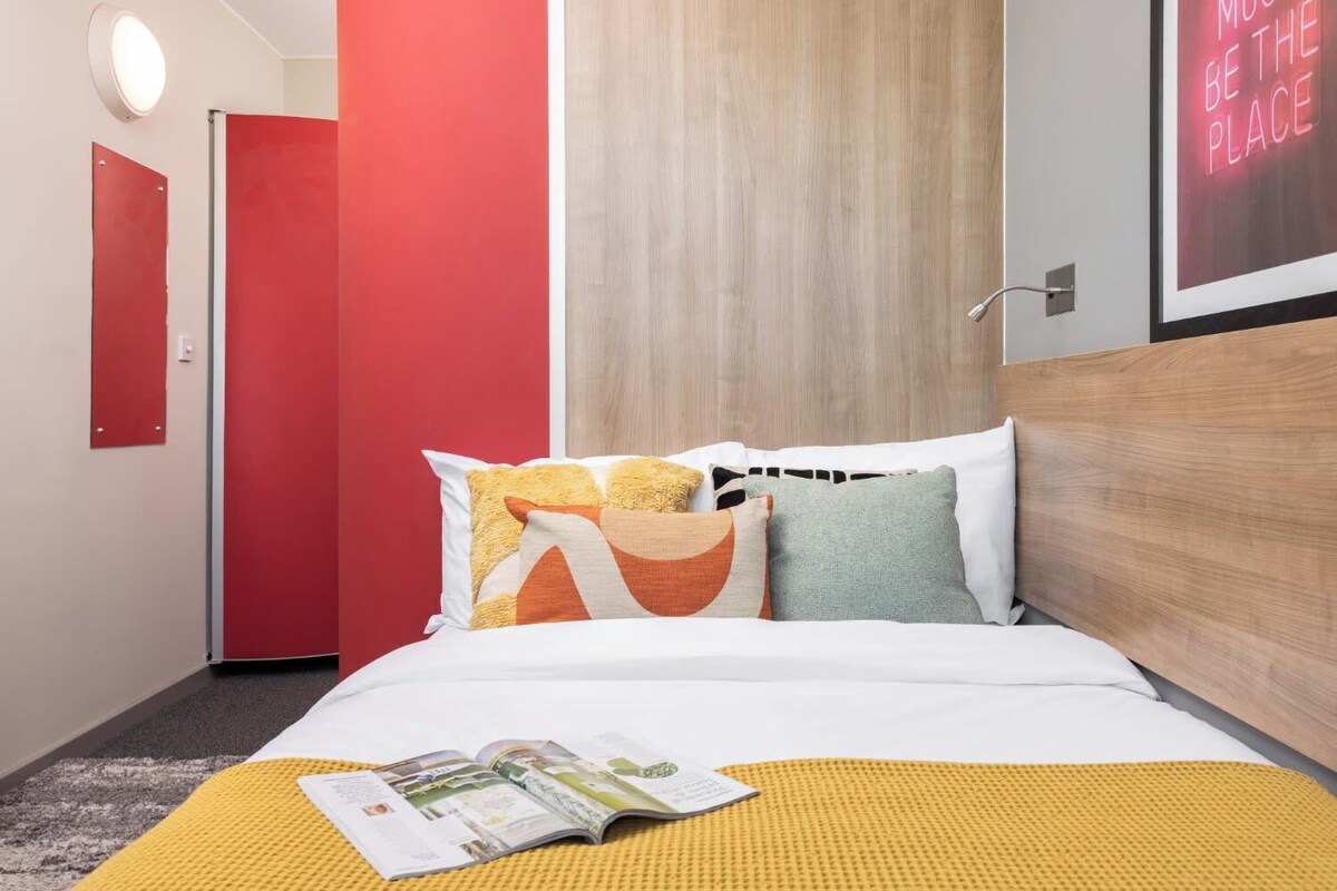 A hotel room with a bed, a magazine on the bed, a red door, and a neon sign on the wall.