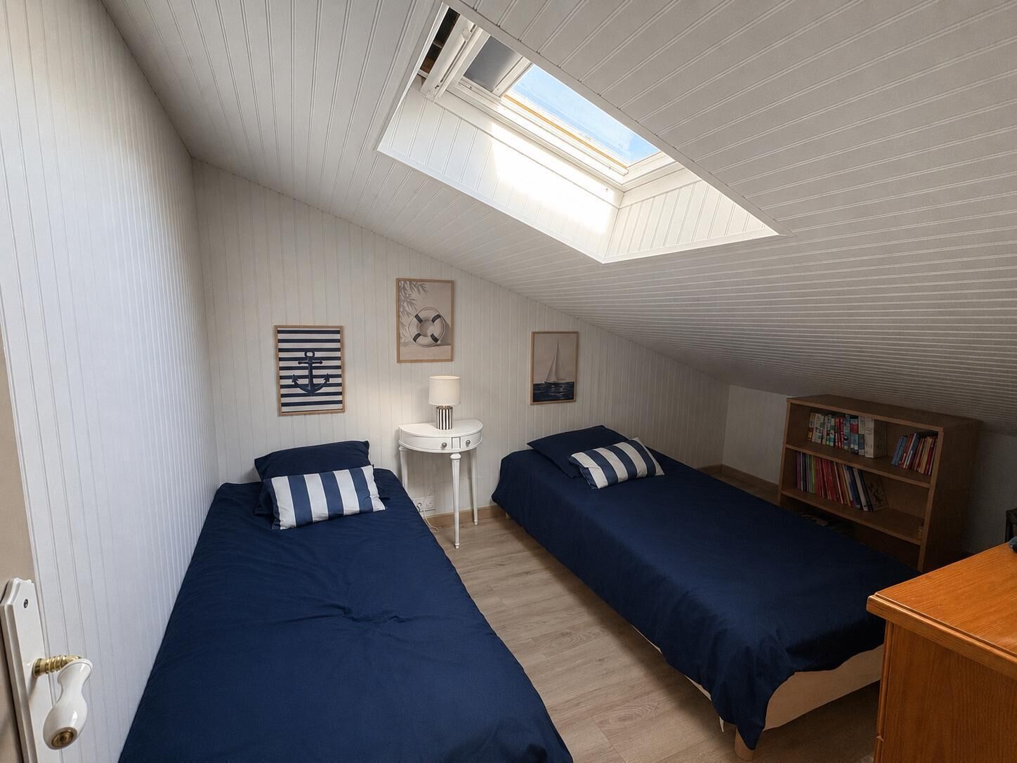 Una habitación con dos camas, una mesita, una estantería y un tragaluz.
