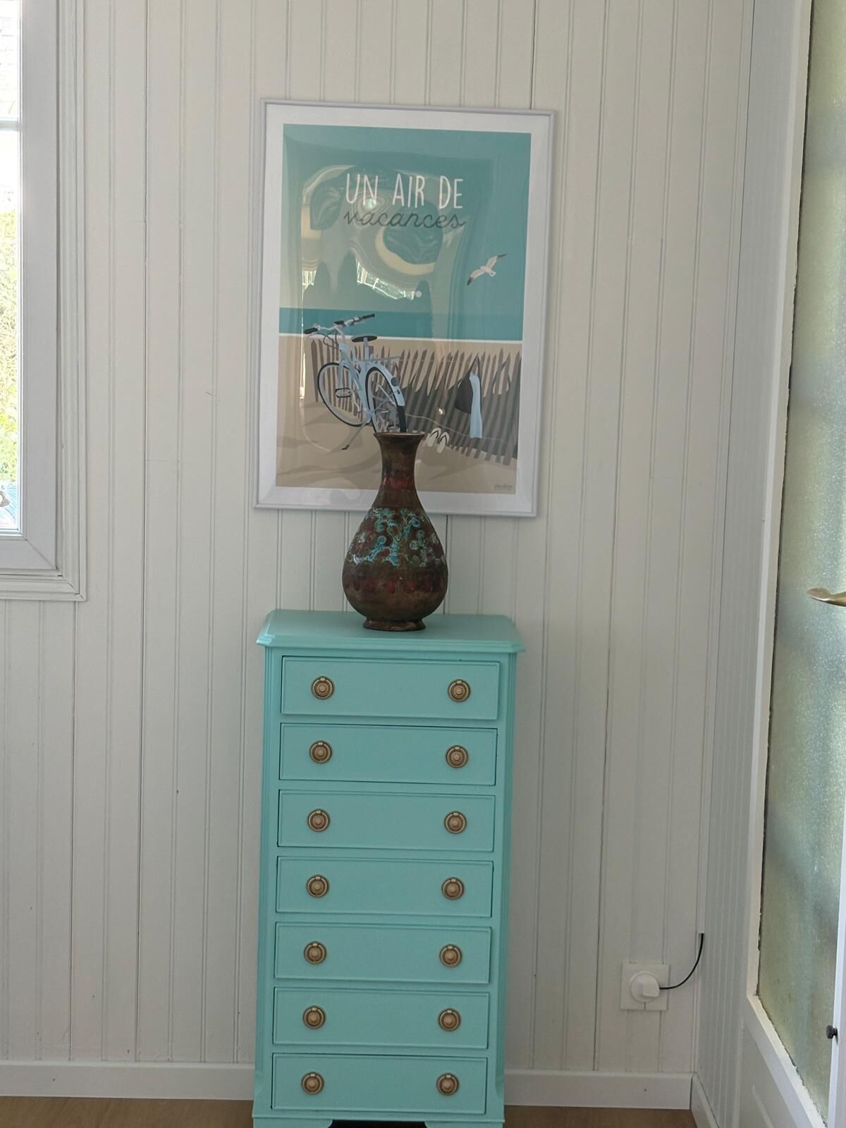 Een smaragdgroene dressoir met gouden knoppen, een decoratieve vaas en een kunstwerk in een lijst op een witte paneelwand.