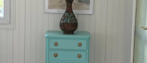 Een smaragdgroene dressoir met gouden knoppen, een decoratieve vaas en een kunstwerk in een lijst op een witte paneelwand.