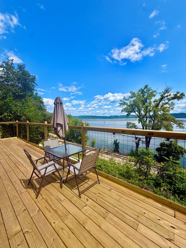 A wooden deck with a glass table and chairs, overlooking a body of water and trees.