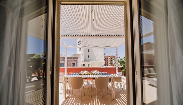 Puerta abierta que da a un patio exterior con mesa y sillas, una pérgola y vistas de edificios al fondo.