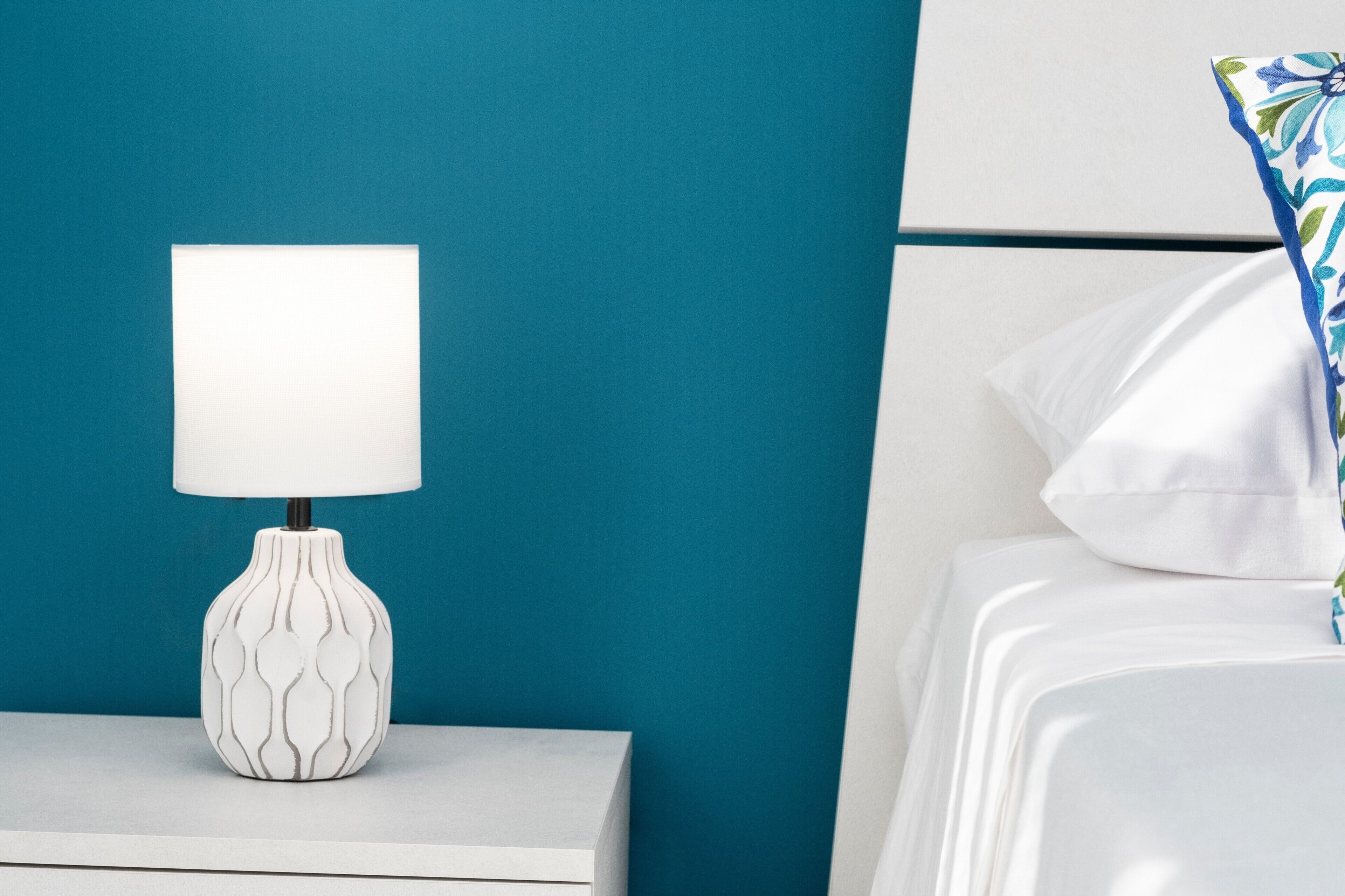 Ein weißer Nachttisch mit einer gemusterten Lampe, eine türkisblaue Wand und ein Bett mit weißer Bettwäsche und einem blumigen Kissen in Blau und Grün.
