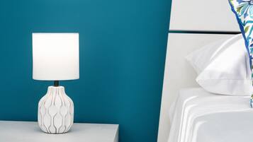 Ein weißer Nachttisch mit einer gemusterten Lampe, eine türkisblaue Wand und ein Bett mit weißer Bettwäsche und einem blumigen Kissen in Blau und Grün.