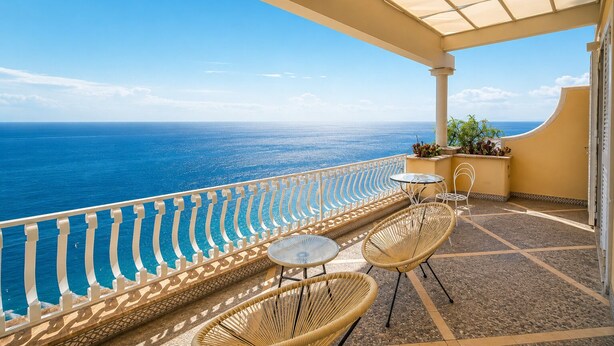 Un balcon avec vue sur l’océan, des fauteuils en osier, une petite table et une plante en pot.