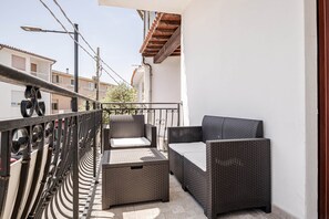 Un balcón con mobiliario de mimbre, baranda metálica y vista a edificios cercanos.
