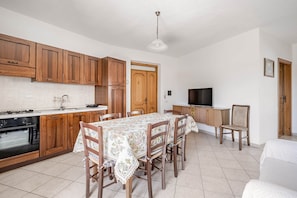 A kitchen with wooden cabinets, a dining table with chairs, a TV, and a sofa. - Domus de Sole | Beach 100m Away & Sea-View Terrace (La Caletta)