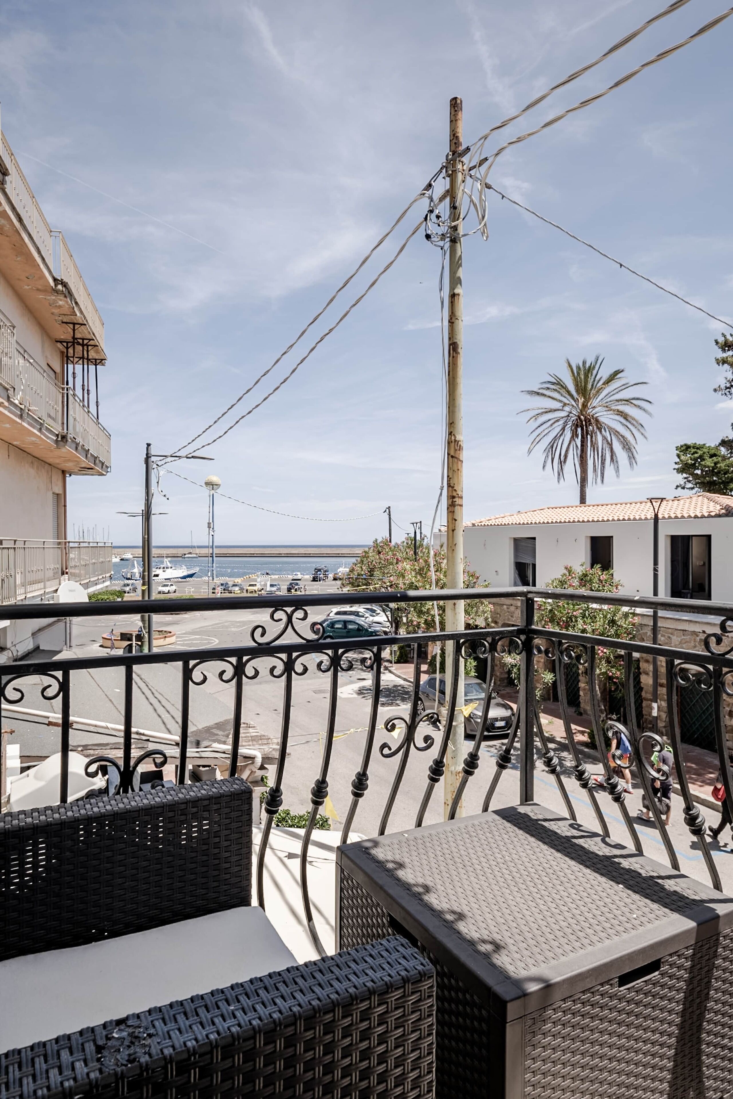A balcony with wicker furniture, a view of the sea, a palm tree, and a building with a tiled roof.