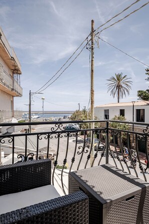 A balcony with wicker furniture, a view of the sea, a palm tree, and a building with a tiled roof. - Domus de Sole | Beach 100m Away & Sea-View Terrace (La Caletta)