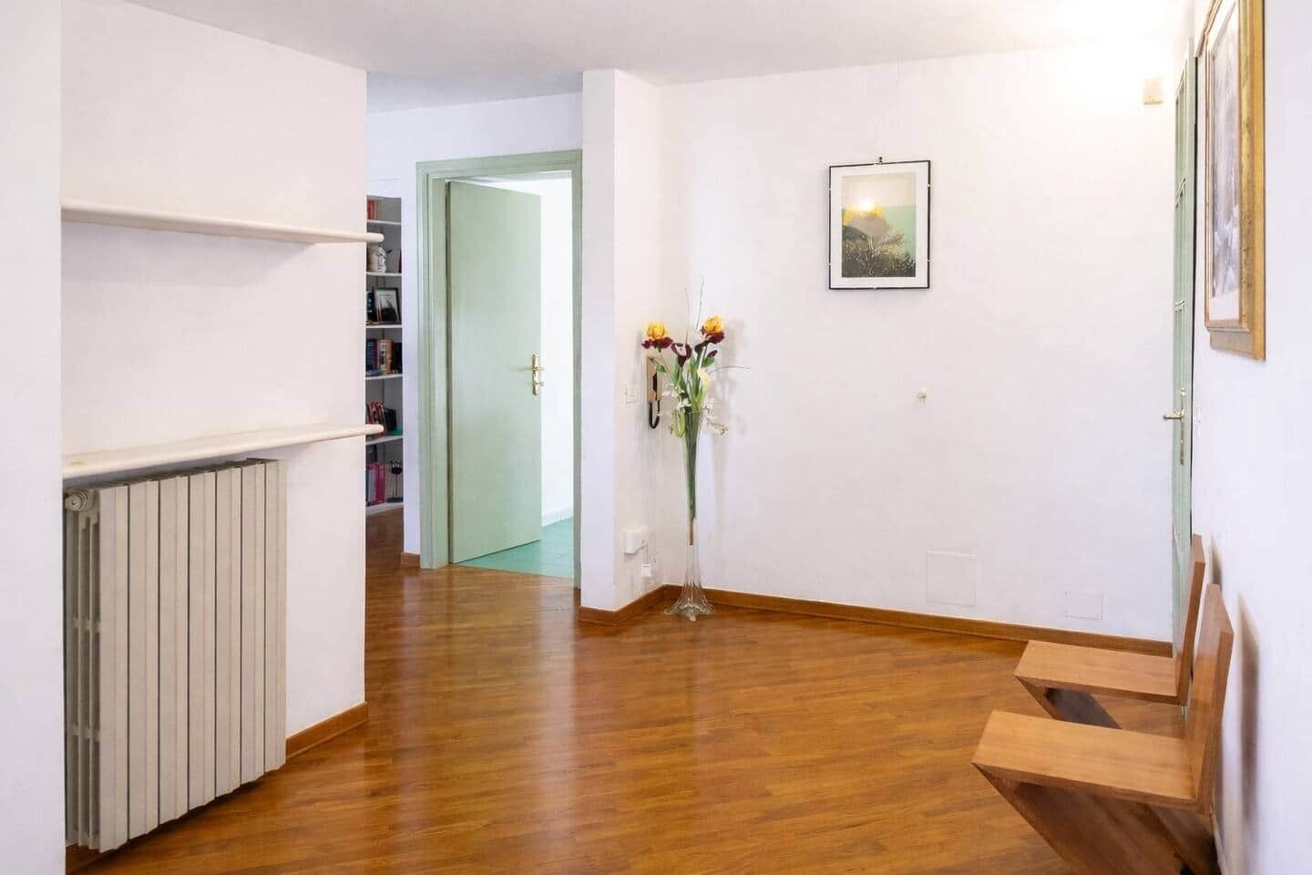 A hallway with wooden flooring, a radiator, a door leading to another room, and a vase with flowers.