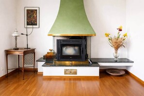 Een houtkachel met een groene kap, een kleine tafel met een lamp, en een vaas met bloemen.
