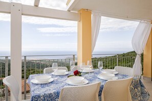 A dining table set for six with white plates and glasses, a blue tablecloth, and a single red rose. The table is on an outdoor balcony with a white railing and a view of the landscape. - Sea View Costa Paradiso | 8 guests (Trinità d'Agultu e Vignola)