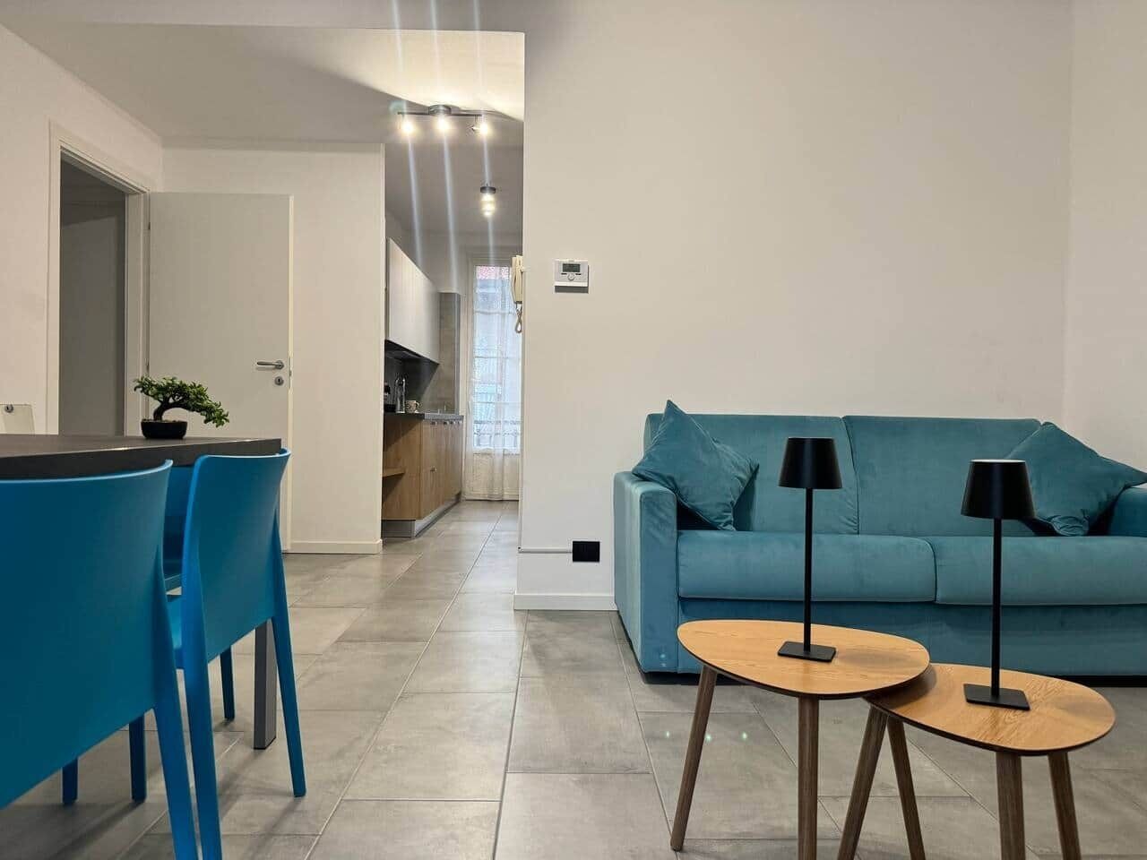 Un soggiorno moderno con un divano blu, due piccoli tavolini e una cucina sullo sfondo.