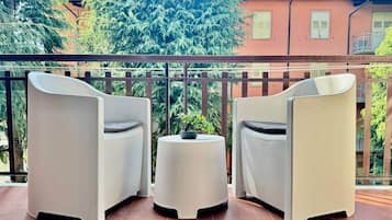 Un balcone con due sedie bianche e un tavolino rotondo, una pianta in vaso e vista su un edificio con tetto rosso.