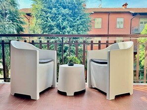 A balcony with two white chairs and a small round table, a potted plant, and a view of a building with a red roof. - Design House | Parking,Terrace (Bergamo)