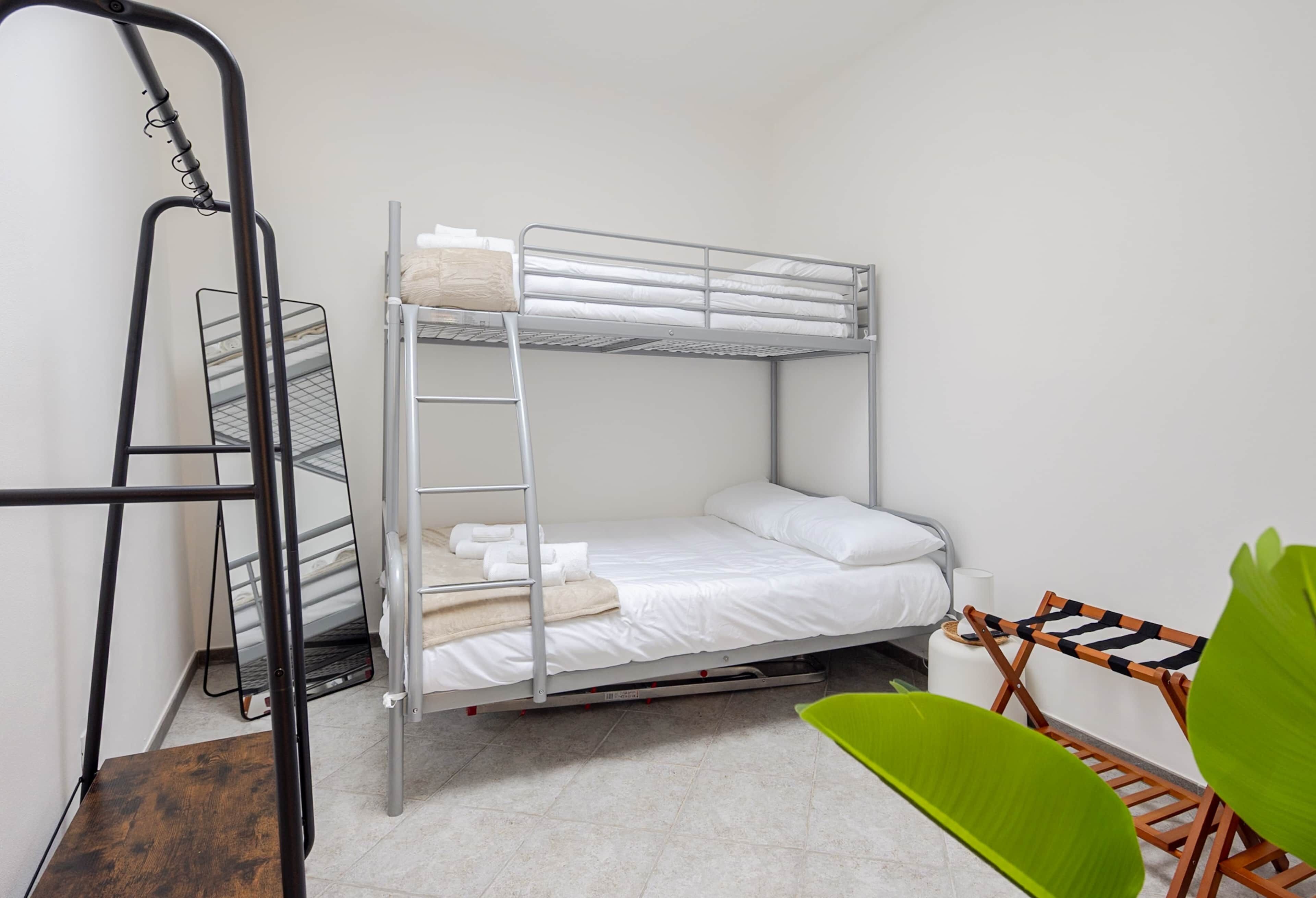 Un lit superposé avec une échelle, une étagère en métal, une chaise verte et un mur blanc.