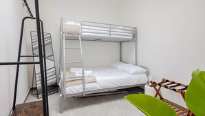 Una cama litera con escalera, un estante de metal, una silla verde y una pared blanca.