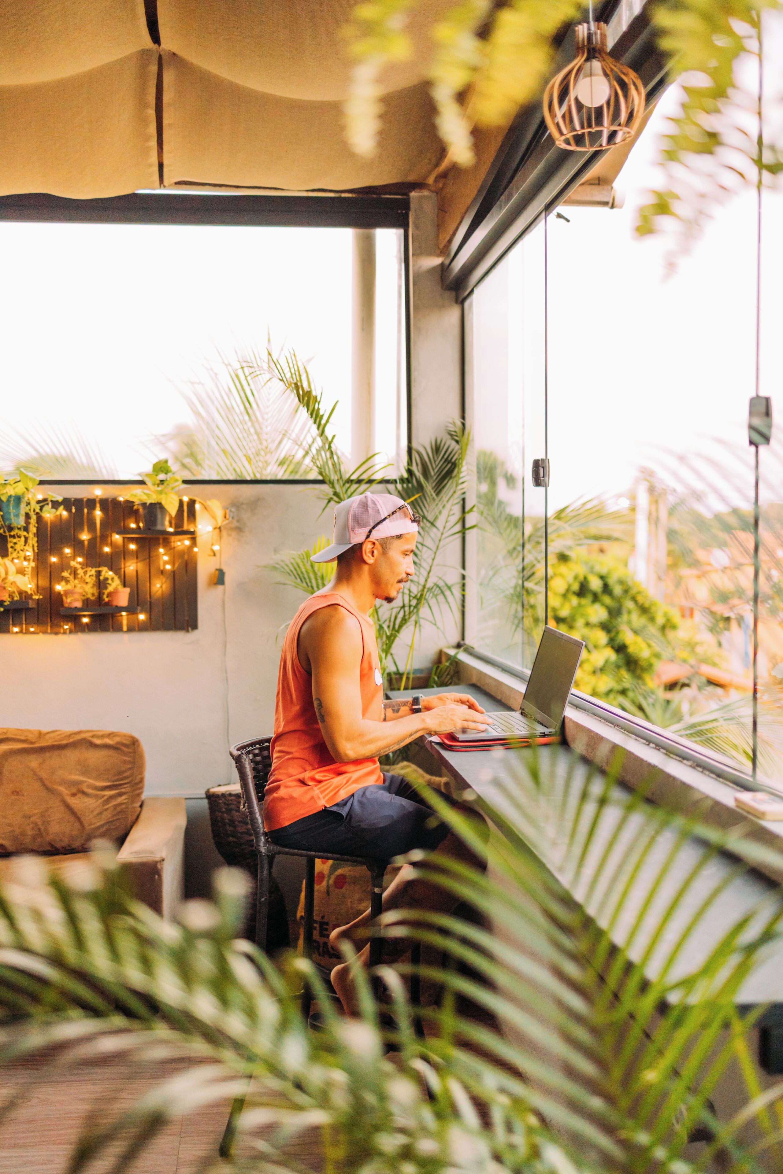 A person seated at a table with a laptop, in a room with large windows, plants, and a hanging light fixture.