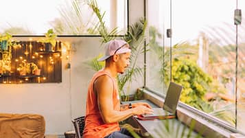 Una persona sentada a una mesa con una computadora portátil, en una habitación con ventanas amplias, plantas y una lámpara colgante.