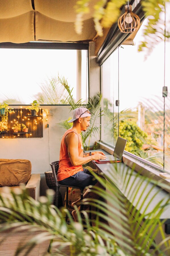 A person seated at a table with a laptop, in a room with large windows, plants, and a hanging light fixture.