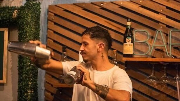 Un barman prépare un cocktail derrière un comptoir, avec un mur fait de lattes de bois et un toit de chaume.