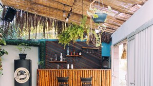 Une terrasse extérieure avec une table en bois, deux tabourets blancs, une plante suspendue, un coin bar avec des bouteilles et des verres, et un toit de chaume.