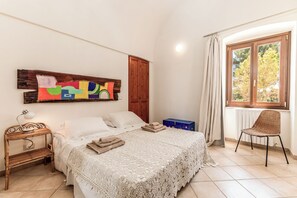 Un dormitorio con cama, mesita de noche, lámpara, silla, ventana con cortinas y armario de madera.