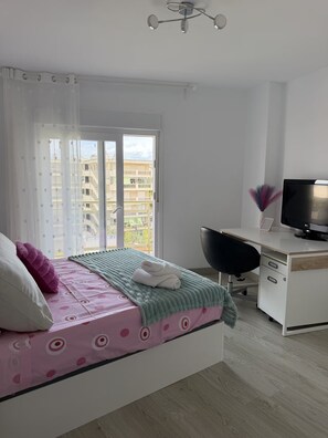 Ein Schlafzimmer mit Bett, Schreibtisch, Stuhl, Fernseher und Balkonblick.