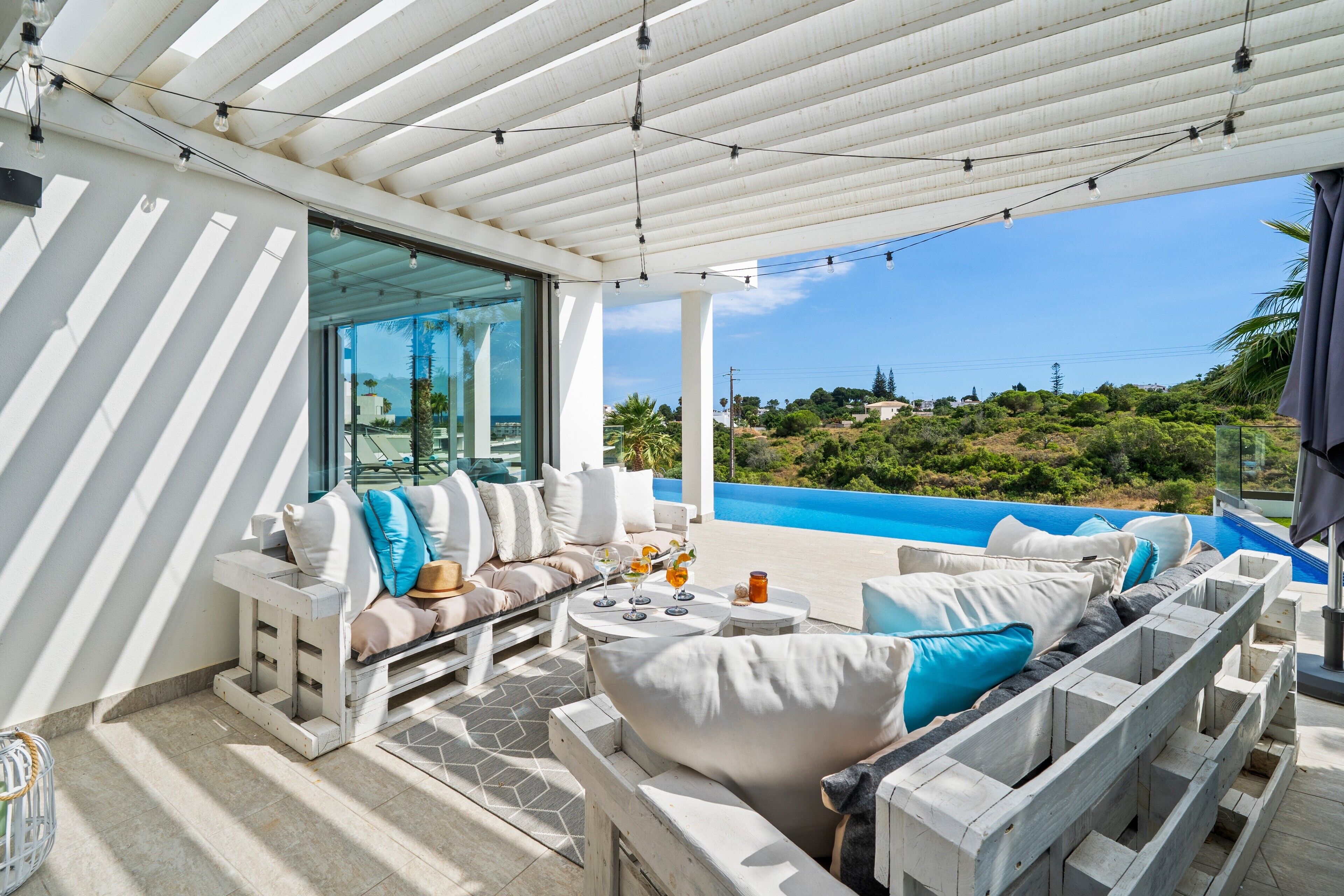 A covered outdoor seating area with a pool view, a sofa with cushions, and a small table with drinks.