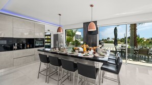 A modern kitchen with a central island, bar stools, and a dining area with outdoor seating. - Villa Catalina with Infinity Pool and Sea View (Armação de Pêra)