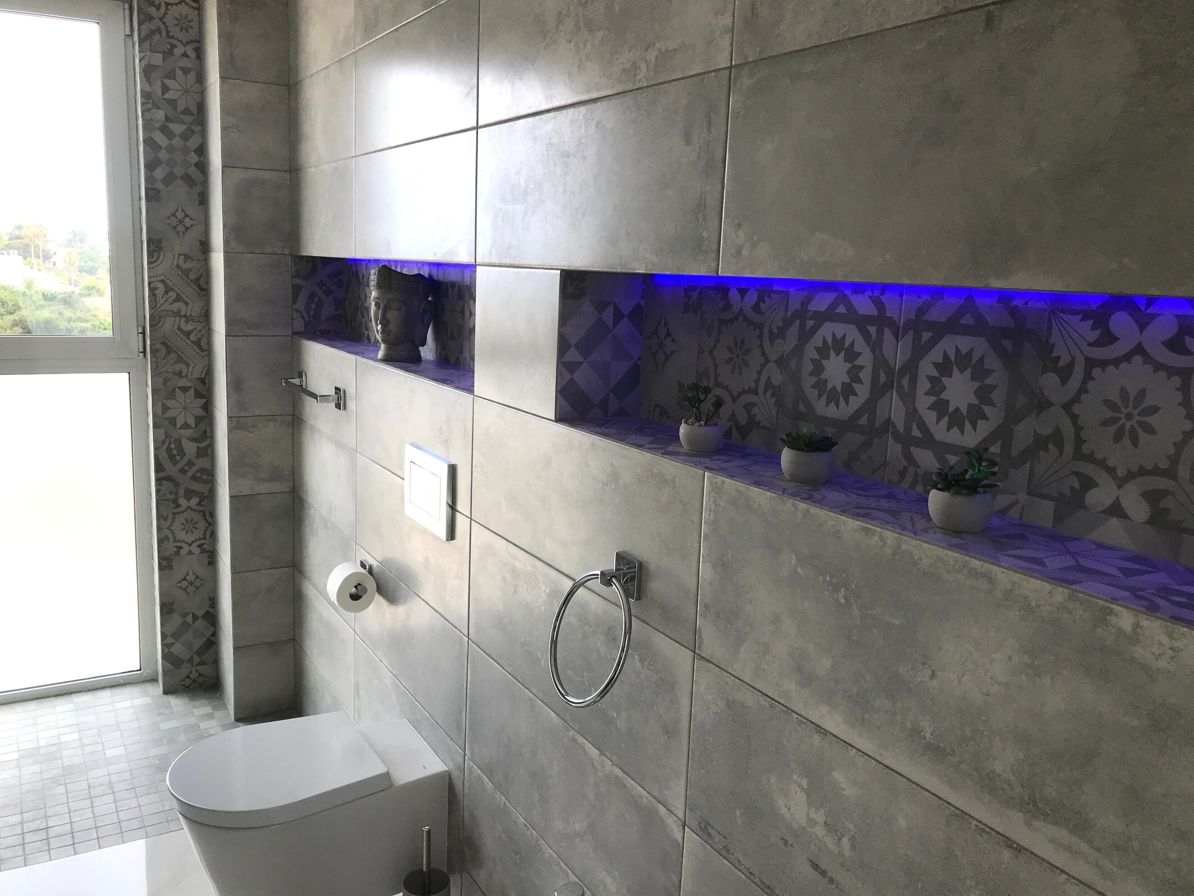 A modern bathroom with grey tiles, a toilet, a bidet, a towel rack, and a shelf with potted plants.