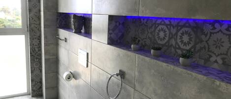 A modern bathroom with grey tiles, a toilet, a bidet, a towel rack, and a shelf with potted plants.