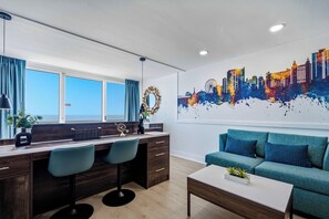 A modern living room with a sofa, a desk with two chairs, a large window, and a cityscape painting on the wall. - Oceanfront Pierfection by Casanita - Lux Suite (Myrtle Beach)