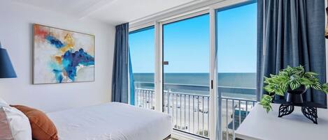 A hotel room with a bed, a painting, a balcony with a view of the beach, and a plant on a table.