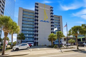 A multi-story hotel building with a sailboat logo, a flag, and a parking lot with cars. - Oceanfront Pierfection by Casanita - Lux Suite (Myrtle Beach)